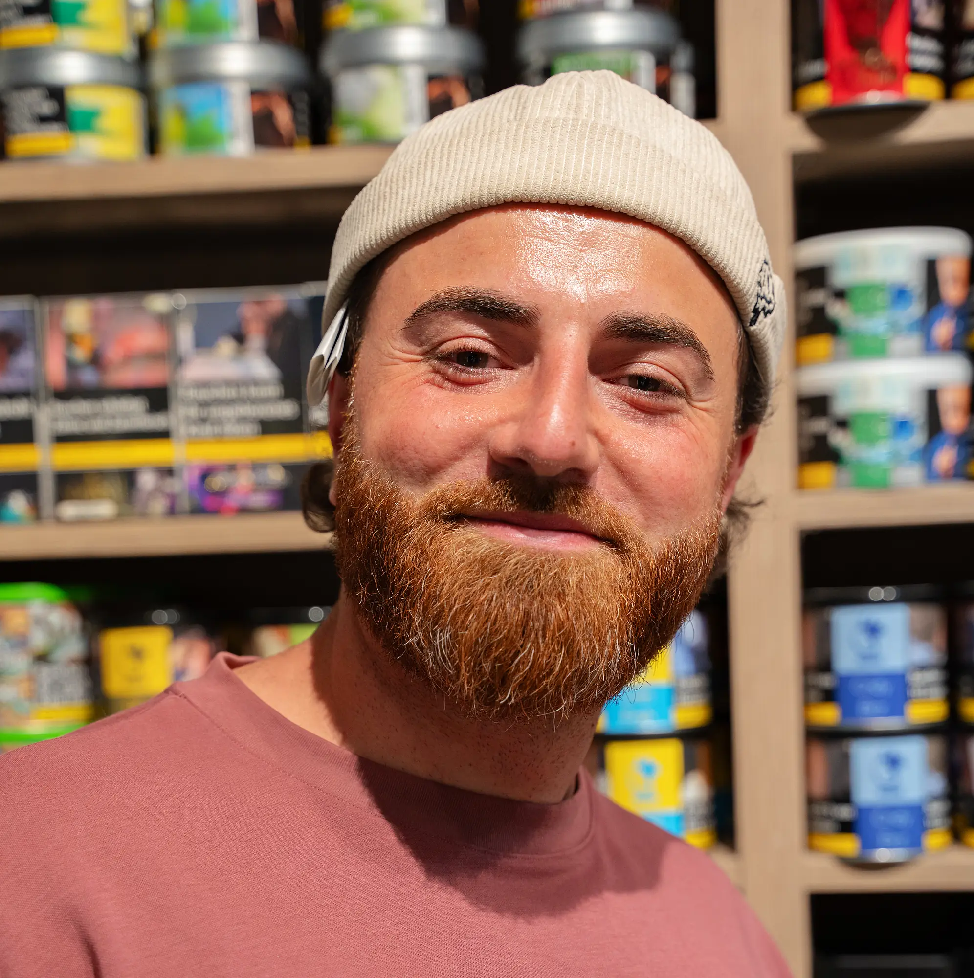 Lächelnder Mitarbeiter der SmokeFactory Köln mit Mütze vor dem Regal mit Shisha-Tabakdosen
