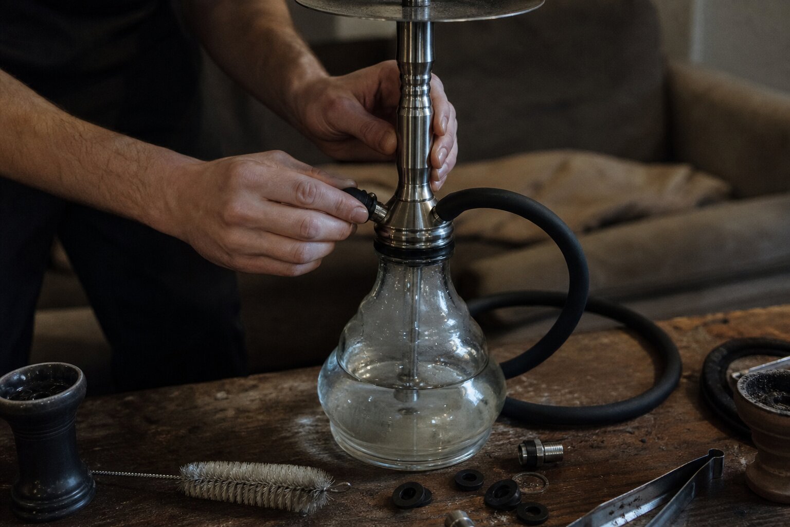 Hände prüfen Schlauchanschluss und Dichtungen einer Shisha auf einem Holztisch, daneben Reinigungsbürste, Gummidichtungen und Kohlezange